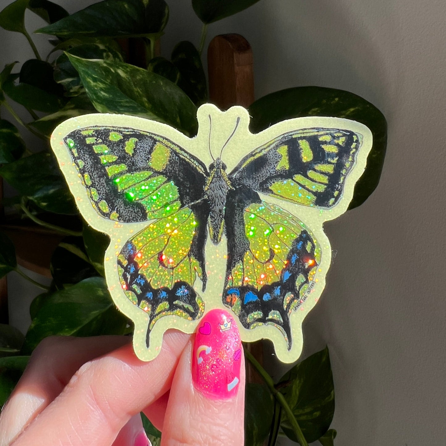 Hand holding a colorful butterfly sticker in front of green leaves